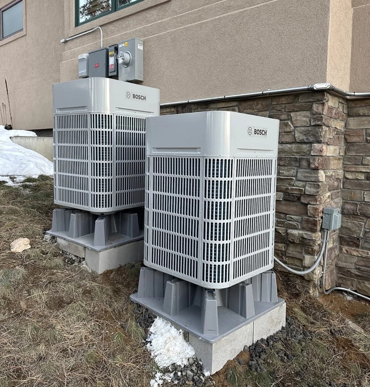 Two Bosch outdoor units on raised bases beside a stone and stucco house