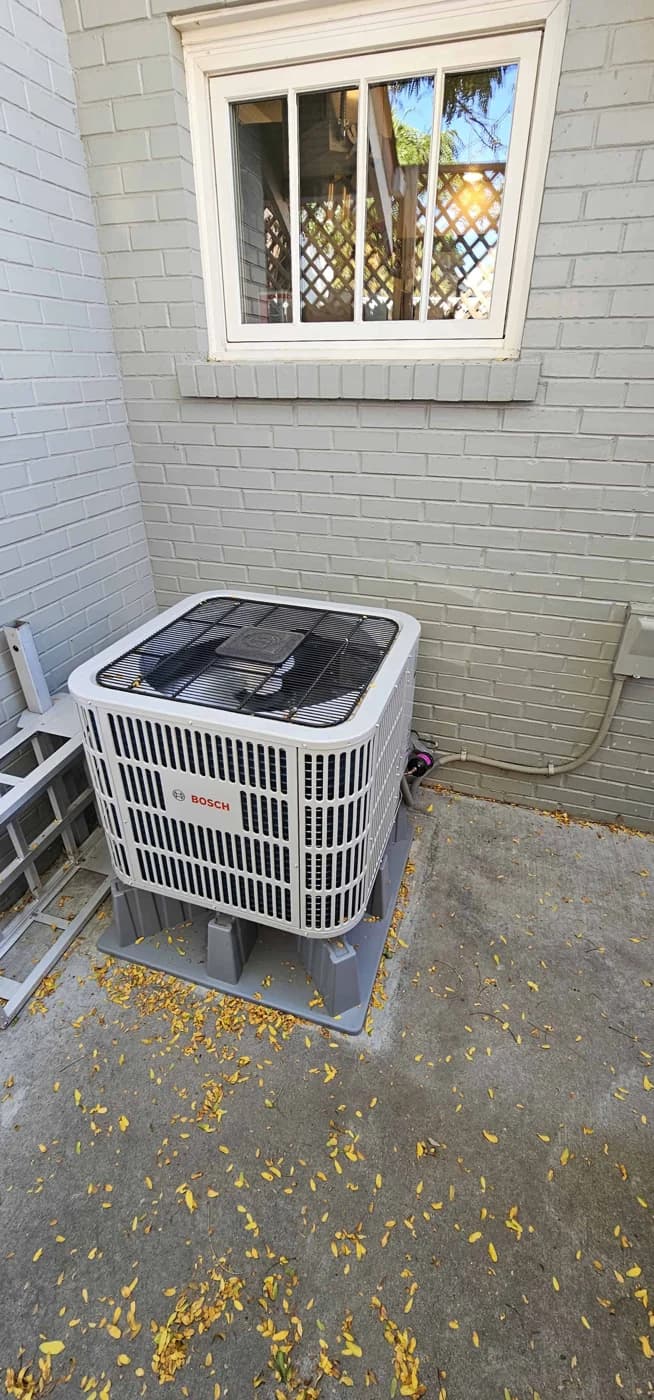 Bosch heat pump beside a gray brick wall beneath a window