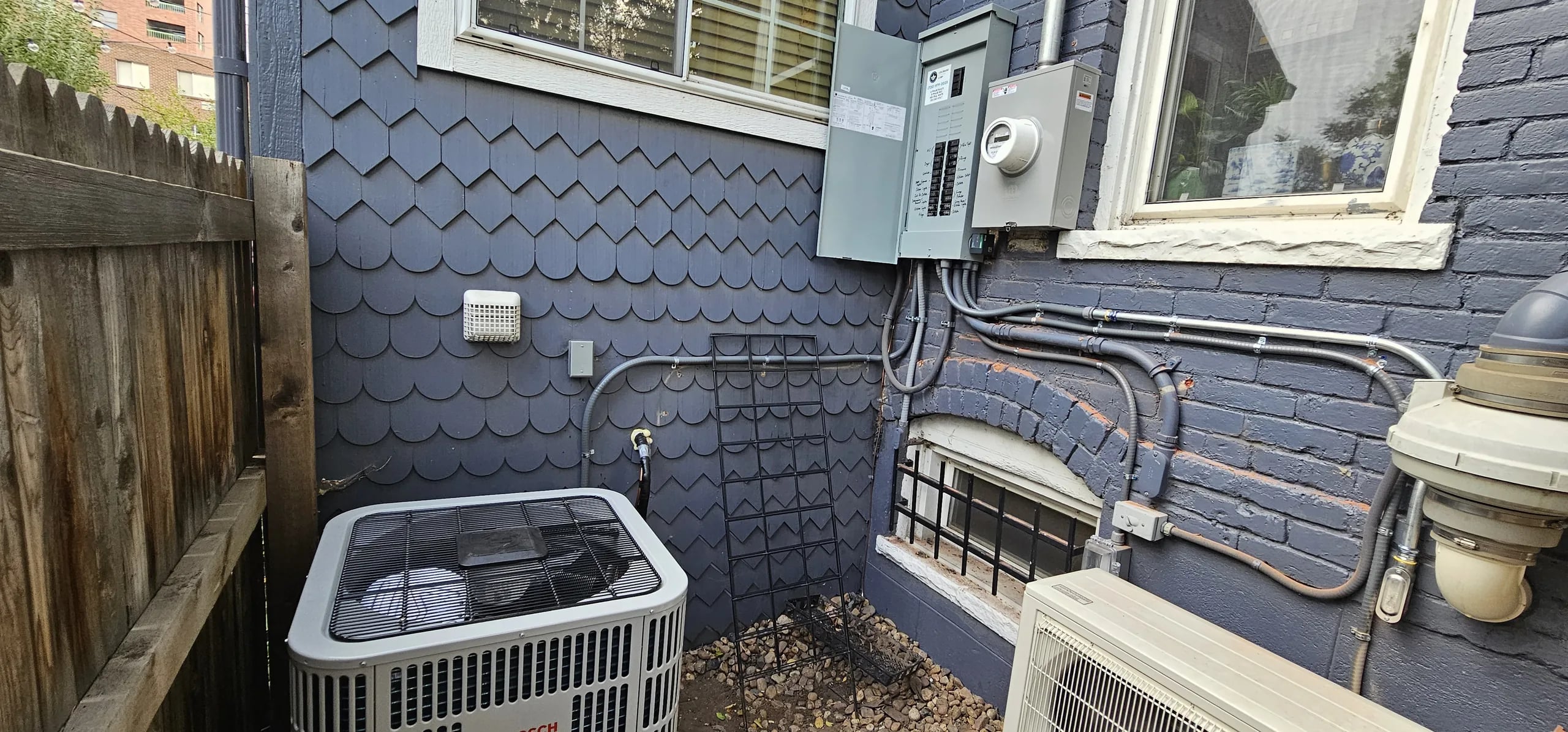 A square outdoor unit and a smaller mini-split outdoor unit in a narrow side yard below the electrical panel on a blue shingled wall