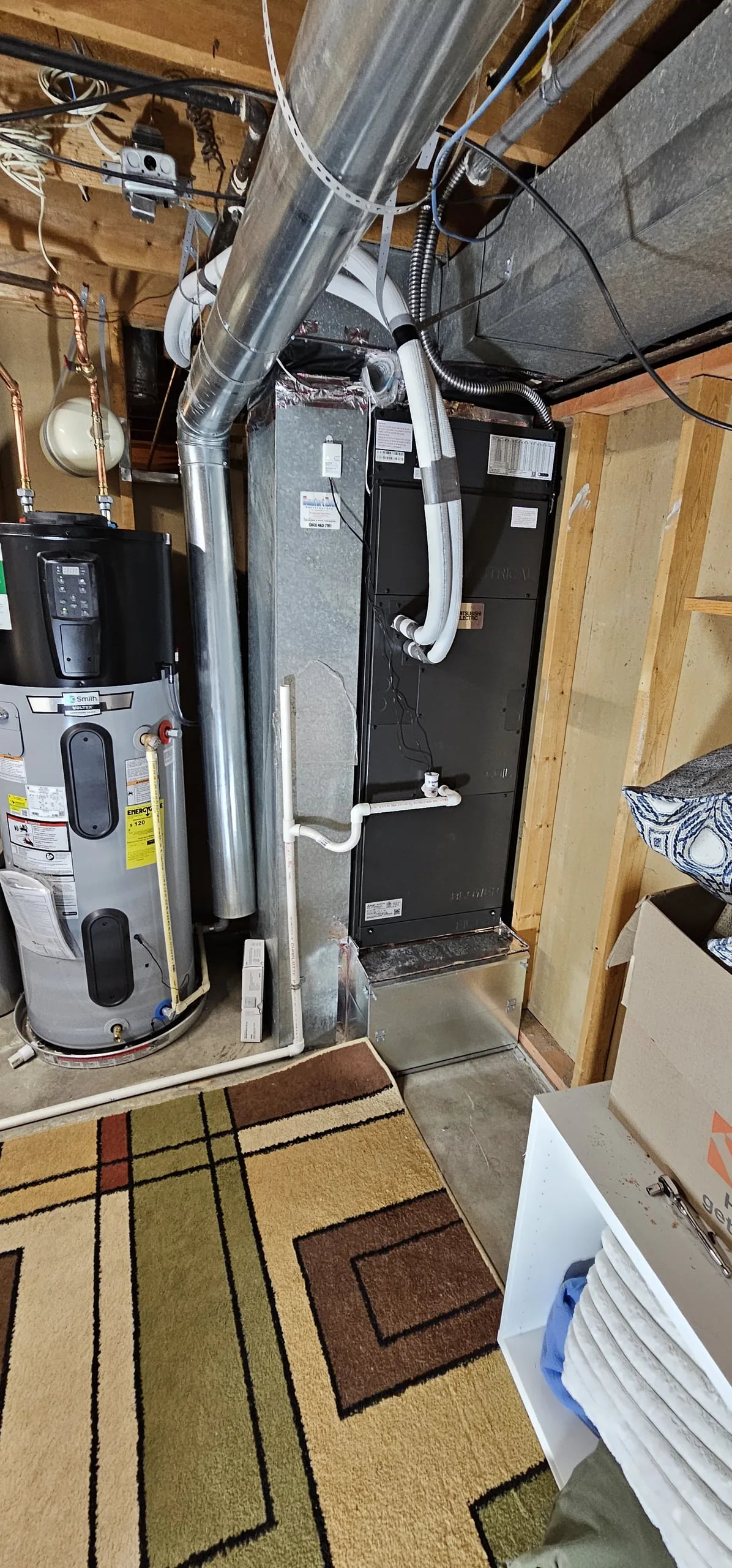 Black air handler with insulated lines running up from its top, beside a water heater in an unfinished basement