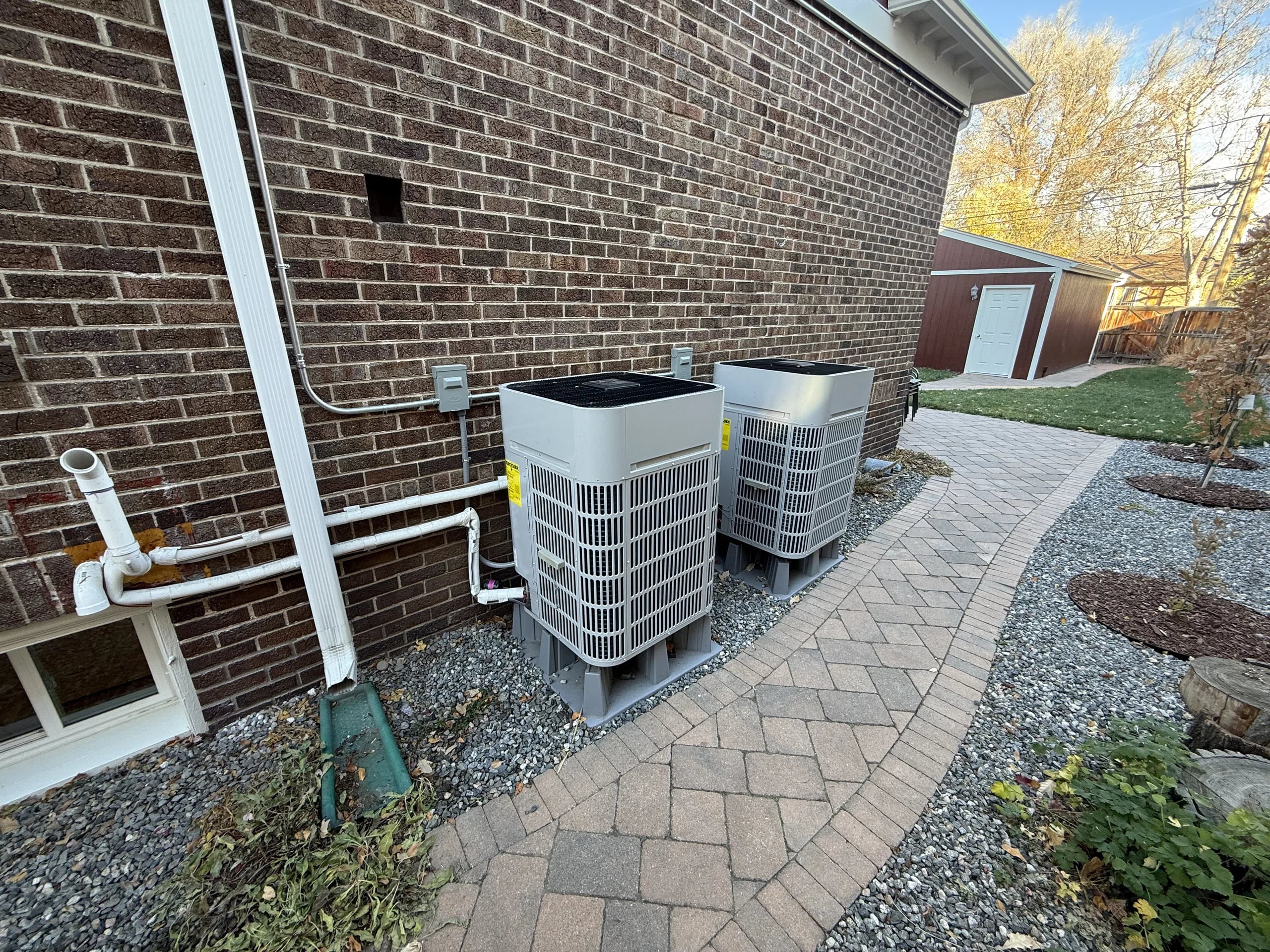 Two outdoor units on risers beside a brick house, with white PVC vent pipes coming out of the wall and a paver path