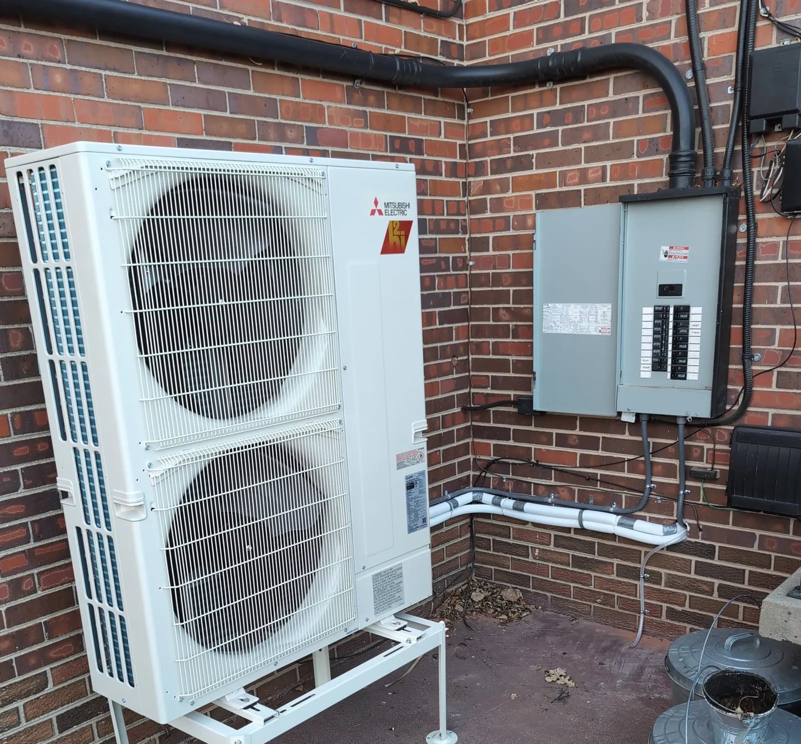 Mitsubishi two-fan outdoor unit on a stand beside a red brick wall, next to the electrical panel and a line-set cover