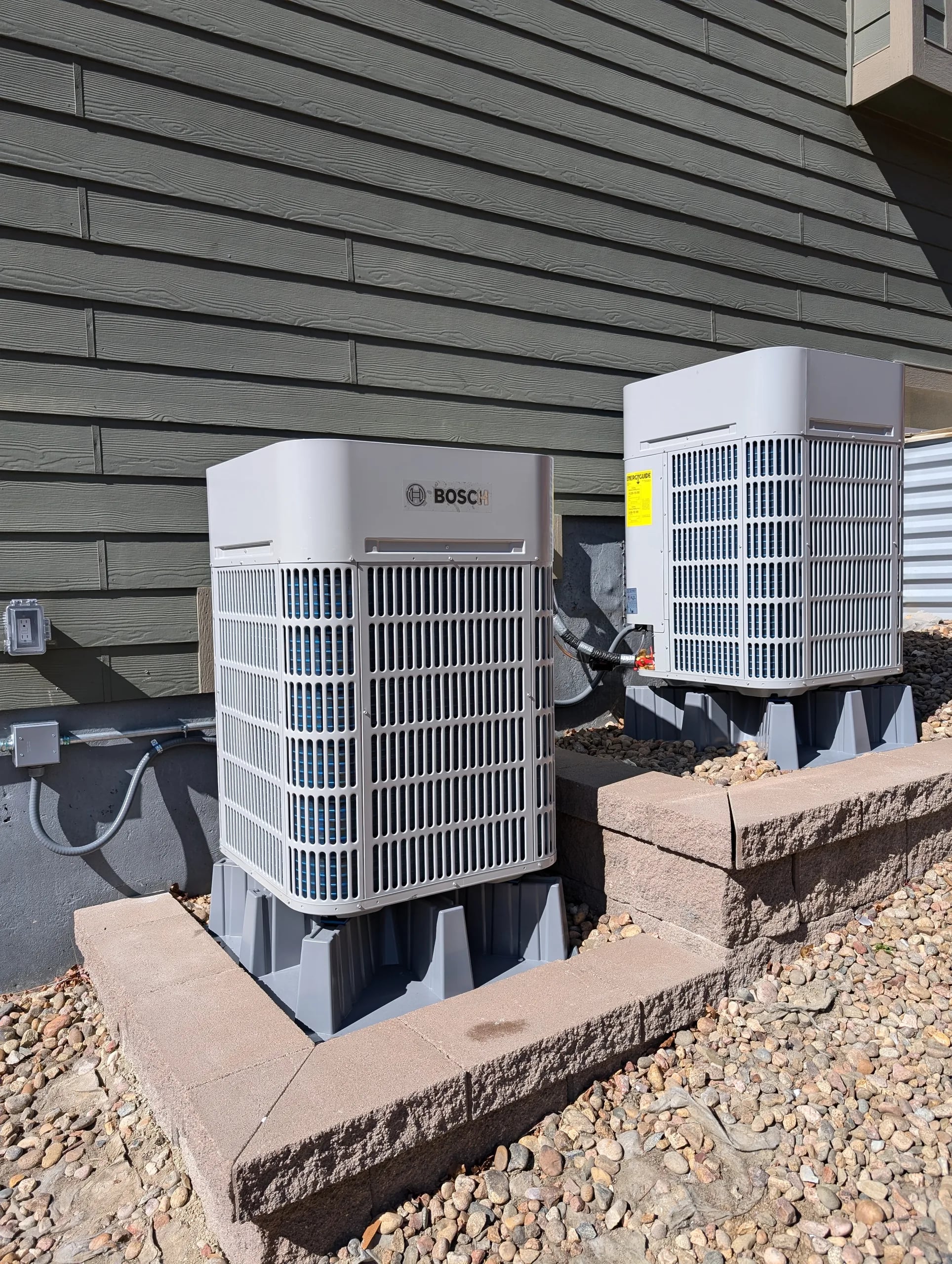 Two Bosch outdoor units on risers on a raised paver pad beside dark gray siding