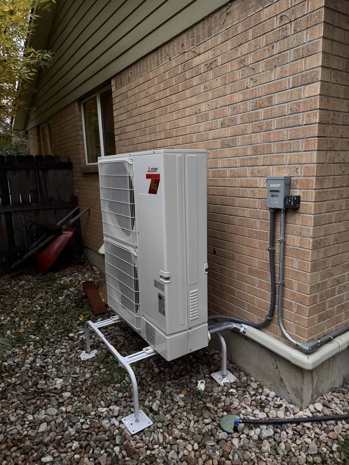 Mitsubishi outdoor unit on a stand on gravel beside a brick wall, with a line-set cover and disconnect